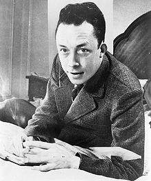 Albert Camus, gagnant de prix Nobel, portrait en buste, posé au bureau, faisant face à gauche, cigarette de tabagisme.jpg